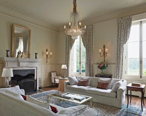 Un salon traditionnel avec une cheminée, un lustre et une grande fenêtre agrémentée de rideaux.