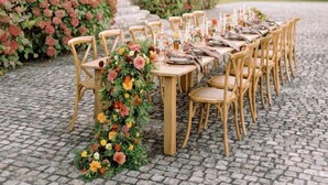 Een lange tafel gedekt voor een maaltijd, met stoelen, een bloemstuk in het midden en een gebouw met een toren op de achtergrond.