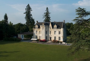 Un grand bâtiment historique, doté de nombreuses fenêtres et d’une façade symétrique, entouré d’une pelouse bien entretenue et de grands arbres.