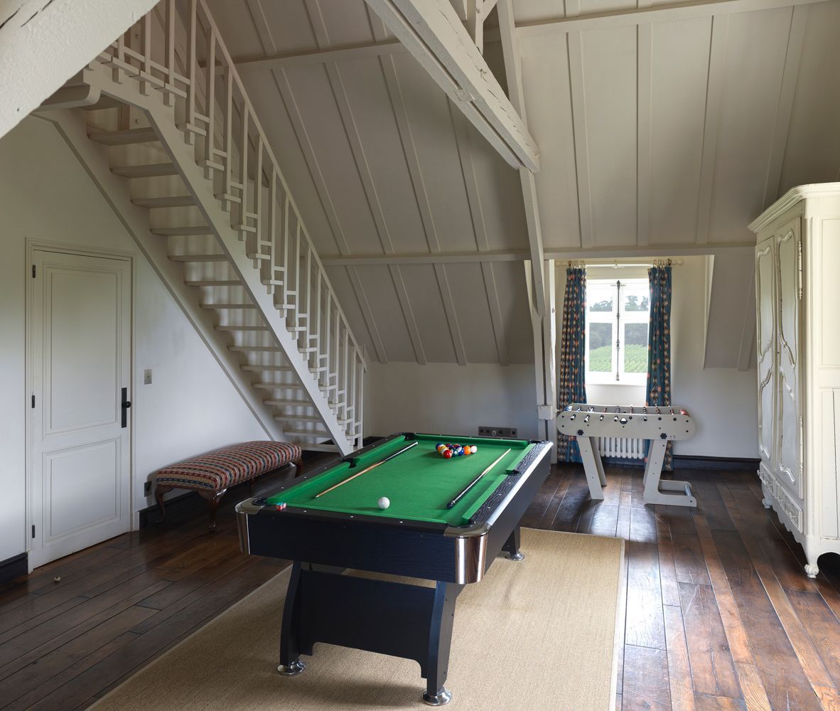 Une table de billard dans une pièce dotée d’un plancher en bois, d’un escalier et d’une fenêtre avec des rideaux.