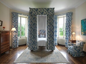Een slaapkamer met een hemelbed, bloemen gordijnen, een houten dressoir en uitzicht op groen.