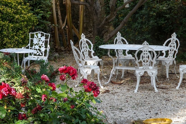A garden with white metal chairs and tables, surrounded by greenery and red flowers.
