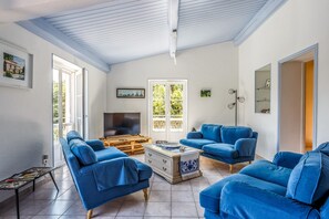 A living room with a TV, two blue sofas, a coffee table, and a large window with a view of greenery. - Villa Du Midi - Four Bedroom Villa, Sleeps 8 (Cruzy)