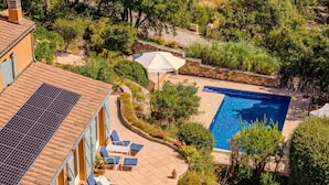 Un área de piscina en la azotea con sillas de descanso y una piscina, rodeada de vegetación y un edificio con techo de tejas.