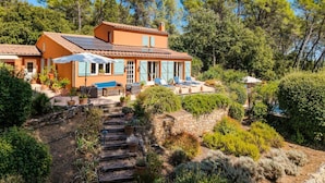 Una villa de estilo mediterráneo con una escalera de piedra que lleva a un patio, rodeado de abundante vegetación y árboles.