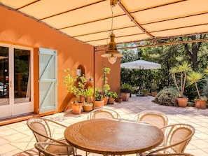 Een terras buiten met een ronde tafel en stoelen, omringd door potplanten en een grote parasol.