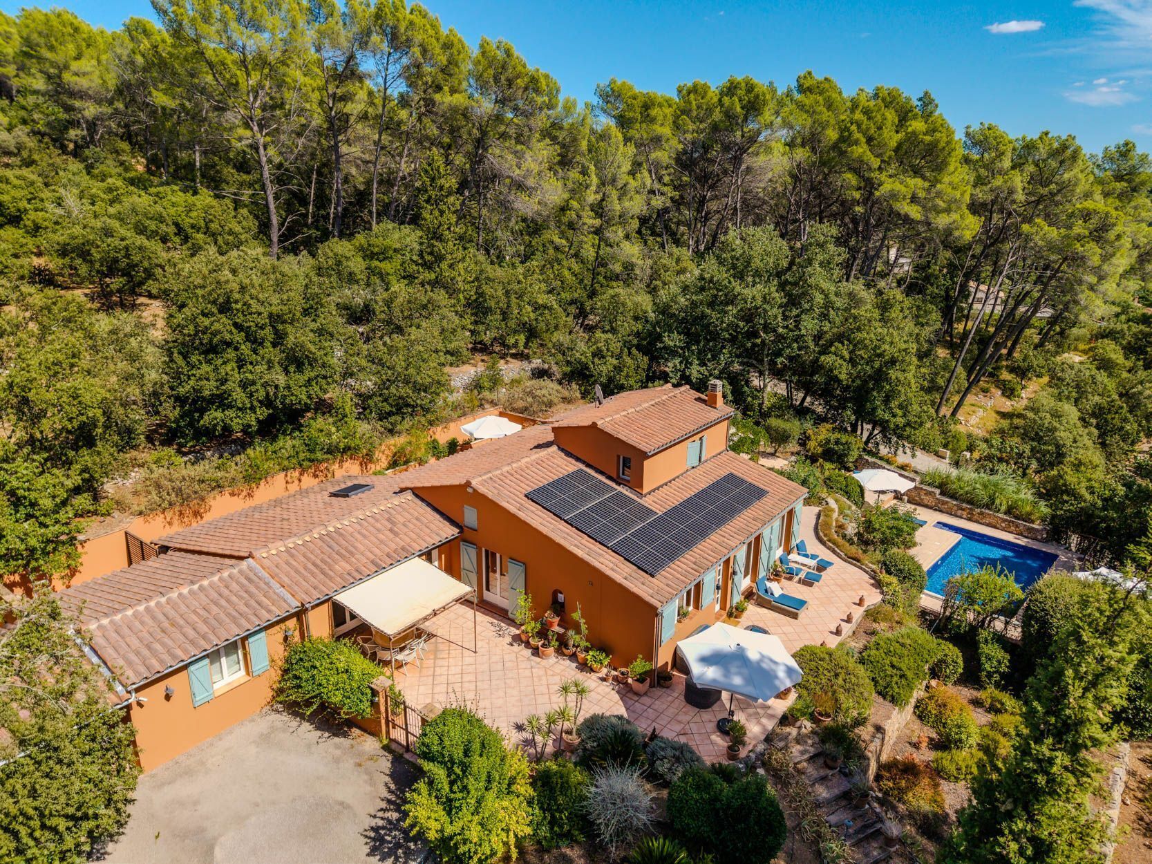 Una villa de estilo mediterráneo con piscina, paneles solares y un patio rodeado de exuberante vegetación.
