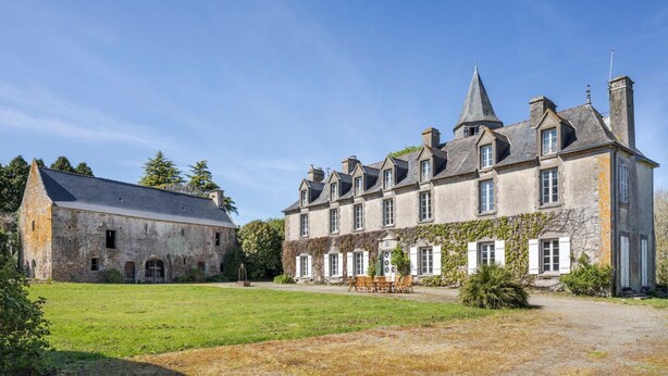 Un grand bâtiment en pierre, doté d’une tour centrale, entouré d’une cour verdoyante et flanqué d’un bâtiment plus petit sur la gauche.