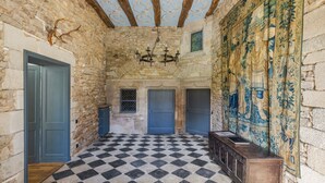 Une pièce aux murs de pierre, au sol à carreaux et ornée d’une tapisserie aux motifs bleus et dorés.