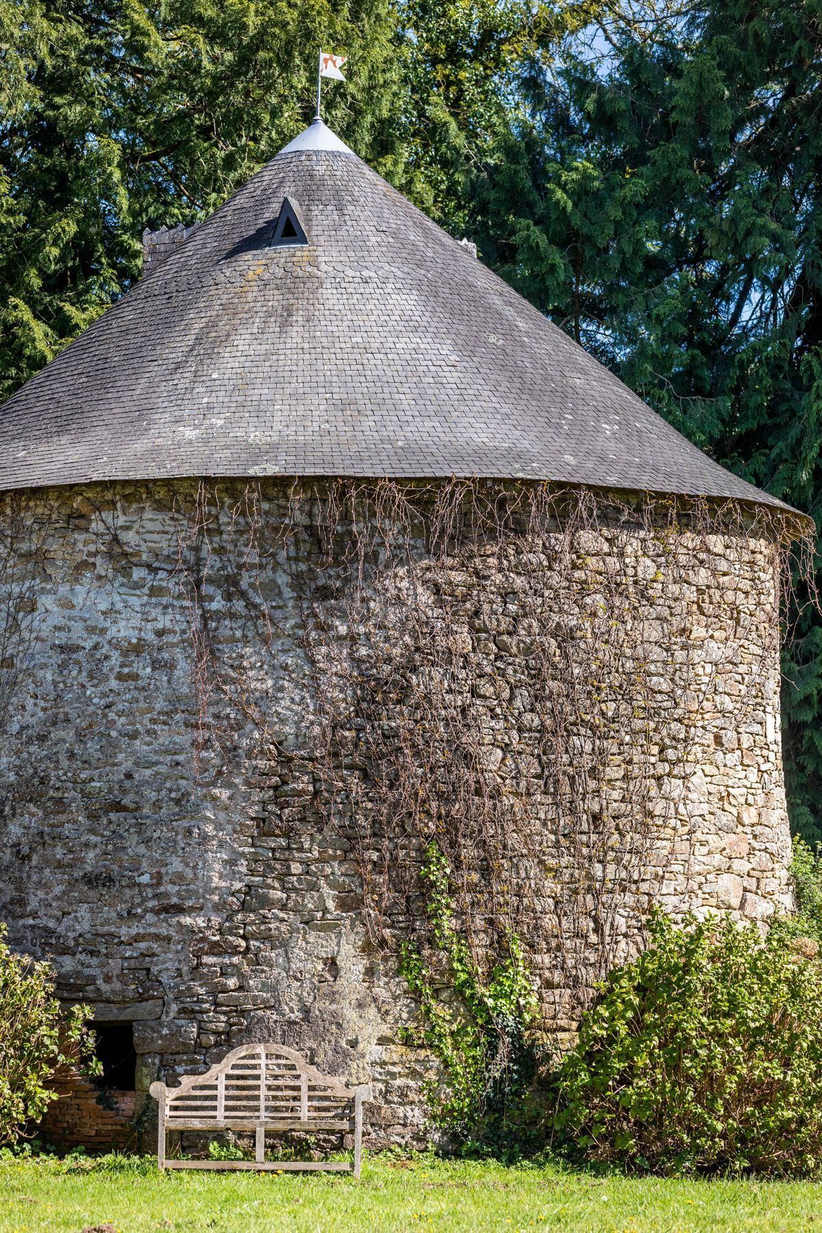 A stone building with a conical roof, a flag on top, and a bench in front.