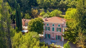A two-story house with a red-tiled roof, surrounded by lush greenery and trees.