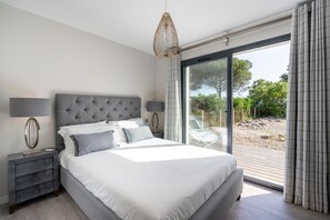 A bedroom with a large bed, a nightstand, a lamp, and a view of the outdoors through a sliding glass door. - Maison Du Lac - Four Bedroom Villa, Sleeps 8 (Carcans)