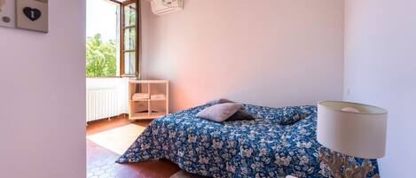Um quarto com cama, tapete redondo, mesa de cabeceira, luminária, janela e radiador.