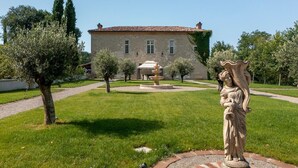 Ein Gebäude aus Stein mit einem Ziegeldach, einer zentralen Fontäne und einer Statue in einem gepflegten Garten.