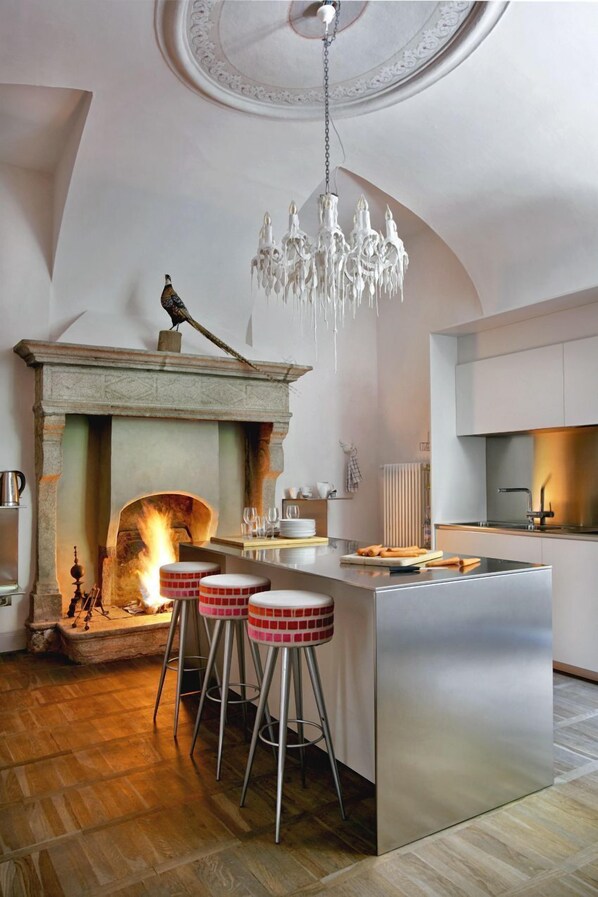 A modern kitchen with a central island, bar stools, a fireplace, and a chandelier.