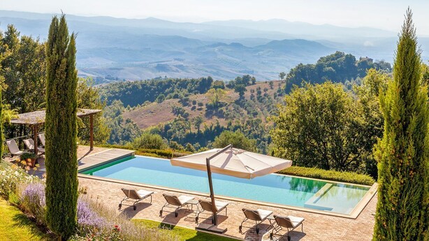 A pool with lounge chairs and an umbrella, surrounded by greenery and a mountainous landscape.