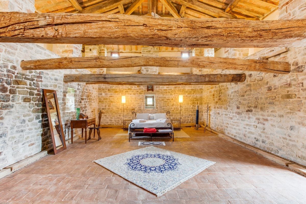 Een rustieke kamer met stenen muren, een houten plafond, een bed, een stoel, een tafel en een vloerkleed.