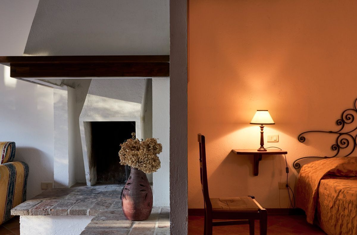 A room with a fireplace, a vase with dried flowers, a chair, a bedside table with a lamp, and a bed with a patterned cover.
