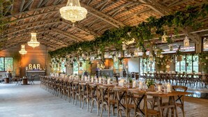 A spacious indoor venue with wooden tables and chairs, chandeliers, hanging lanterns, and a bar area.