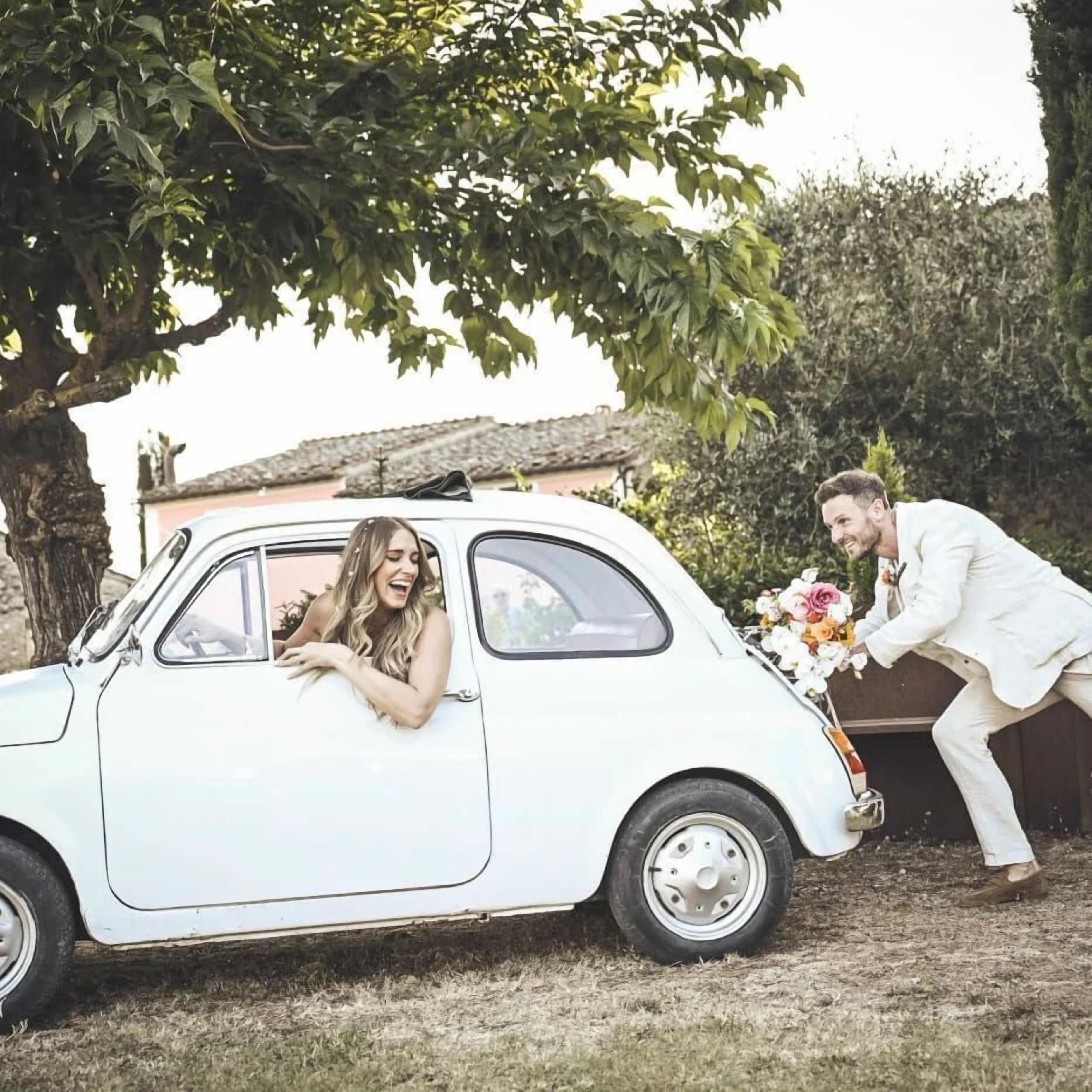 A white car parked under a tree with a person leaning out of the window and another person holding a bouquet of flowers.
