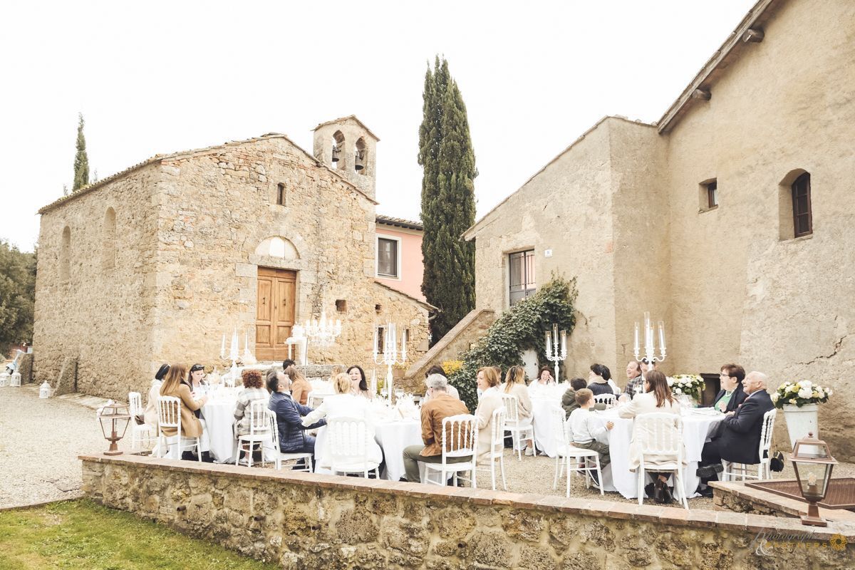 An outdoor wedding reception with guests seated at round tables, stone buildings in the background, and a clear sky.