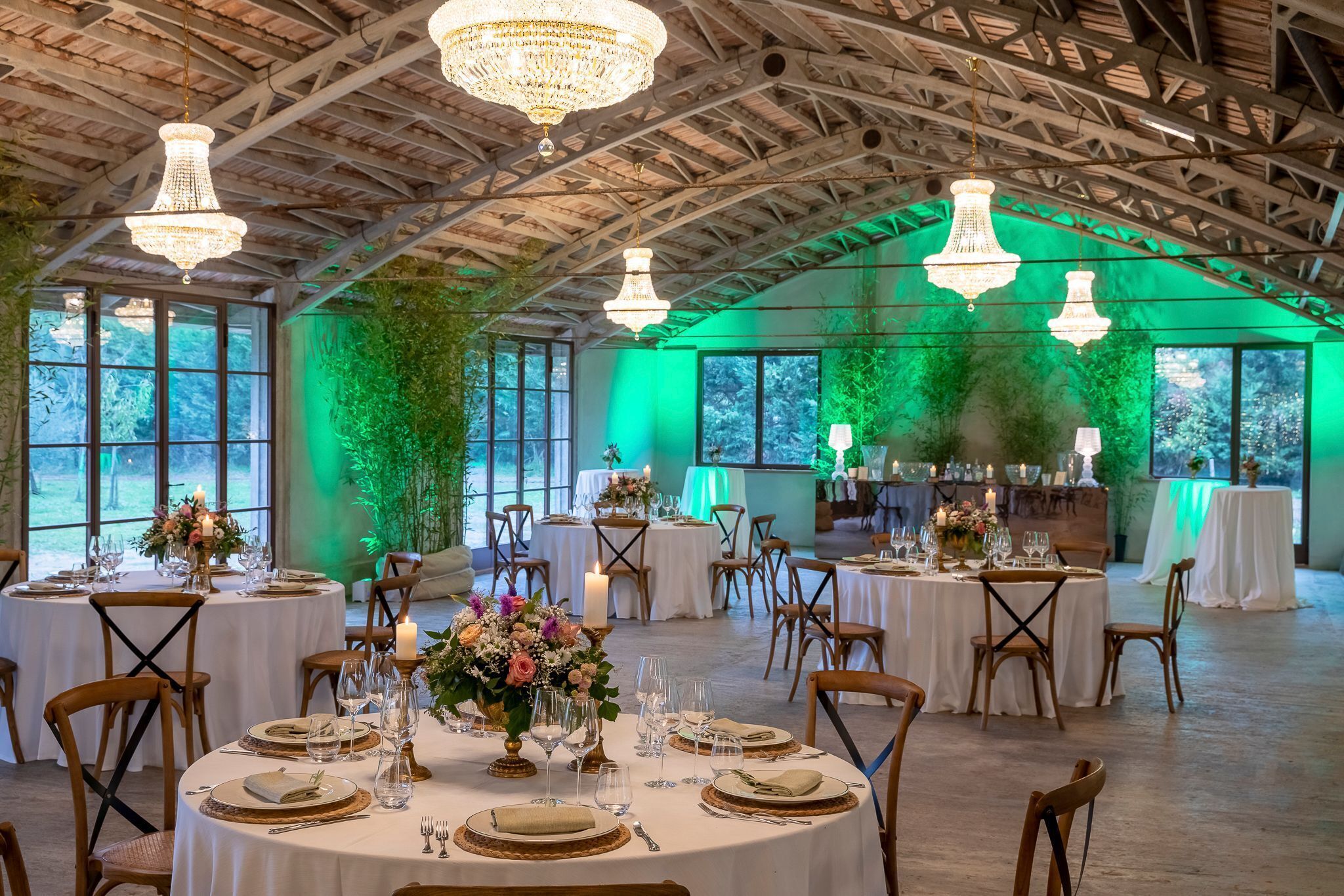 Une salle de banquet avec des tables rondes dressées pour un événement formel, agrémentée de lustres, de compositions florales et d’un plafond en bois rustique.