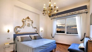 Un dormitorio con una cama grande, una mesita de noche, una silla, una araña y una ventana con cortinas.