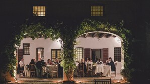 Un espacio de comedor al aire libre con mesas y sillas, rodeado por un jardín y un edificio con ventanas.