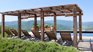 Une pergola en bois, agrémentée de six chaises longues et de plantes en pot, offrant une vue sur un paysage montagneux et une piscine.