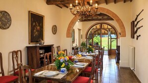 A dining area with a long wooden table set for six, red cushioned chairs, a chandelier, and a view through an arched doorway to the outdoors.