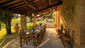 An outdoor covered dining area with stone flooring, wooden tables and chairs, and a view of a vineyard in the background.