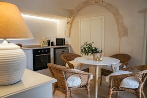 A compact kitchen with a microwave, kettle, and oven. There is a round dining table with four wicker chairs and a potted plant on it. - La Piccola Pietrina - One Bedroom Villa, Sleeps 2 (Ostuni)