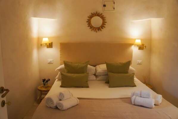 Habitación de hotel con una cama, dos lámparas de pared, un reloj de pared y una mesita con un florero y toallas.