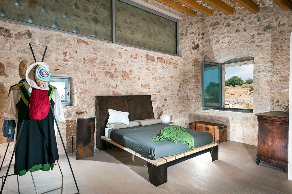 A bedroom with a stone wall, a wooden bed, a window with a view, and a mannequin with a hat.