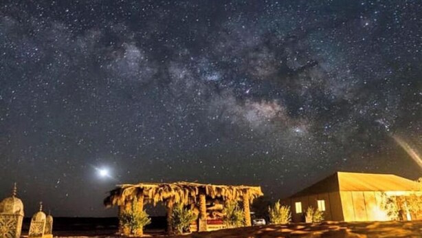 A night scene with a starry sky, a thatched-roof structure, and a building with a sloped roof. - Fabulous Desert Camp (Merzouga)