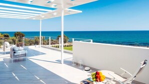 Een ruim balkon met vrij uitzicht op de oceaan, witte meubels en een schaal fruit.