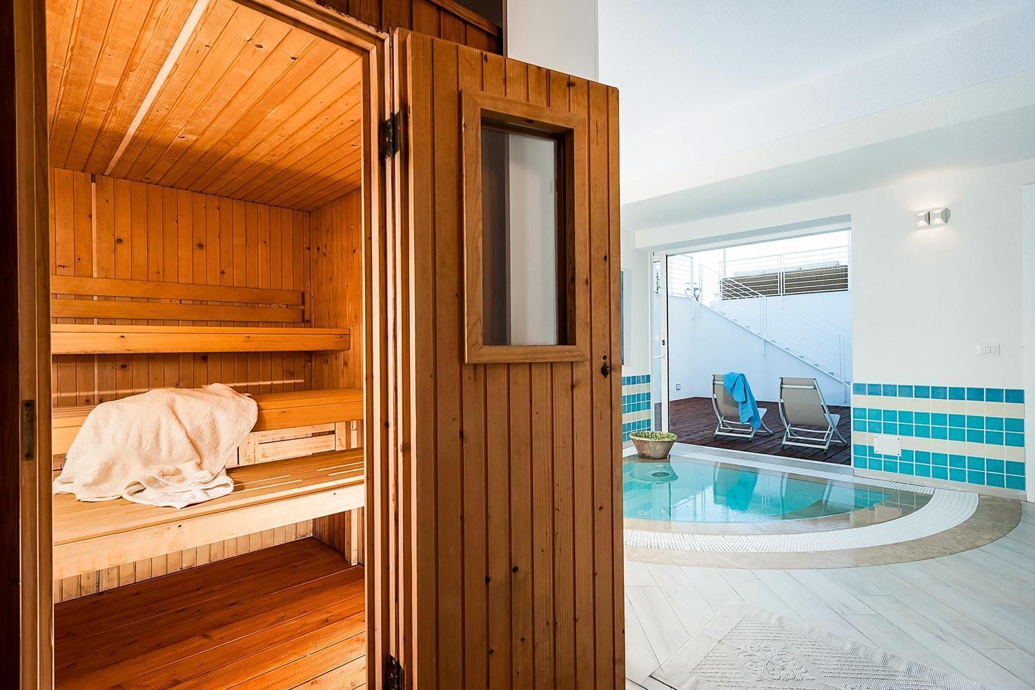 A wooden sauna with benches and a towel, a pool area with lounge chairs, and a tiled wall.
