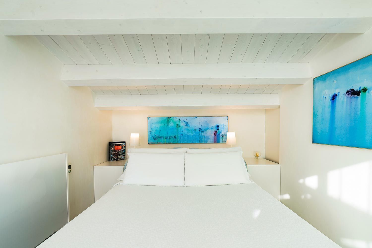 A hotel room with a large bed, two bedside tables, a painting on the wall, and a ceiling with wooden beams.