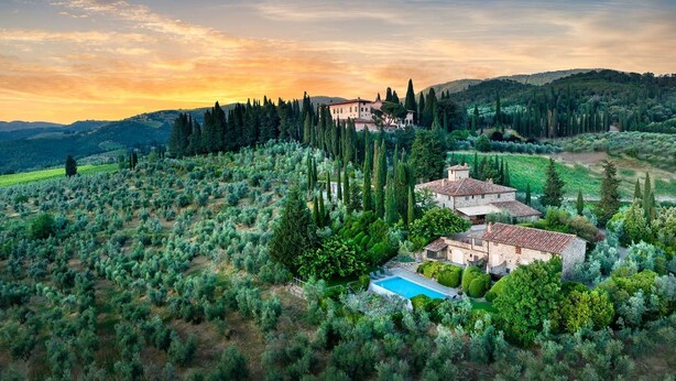 Une villa dotée d’une piscine, entourée d’oliviers et de cyprès, avec en toile de fond des collines ondoyantes et un ciel au coucher du soleil.