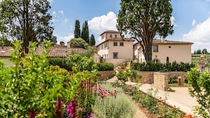 Un jardin où s’épanouit une grande variété de fleurs, avec un sentier qui mène à un bâtiment doté de nombreuses fenêtres et d’un toit recouvert de tuiles.