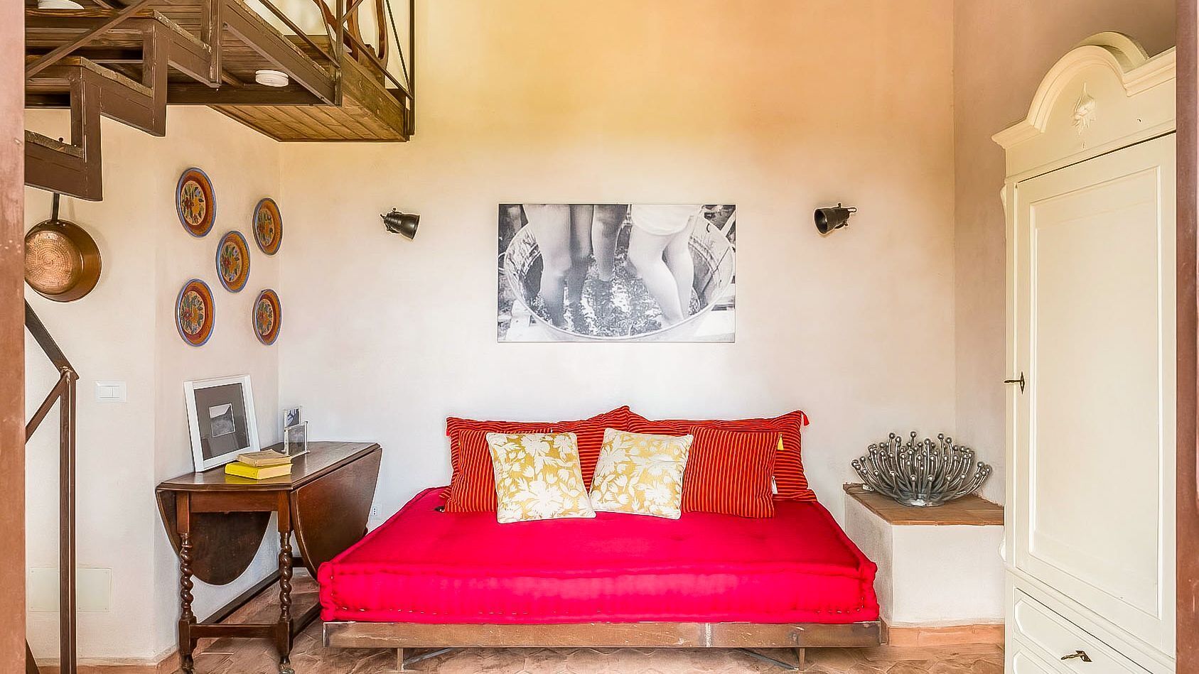 A bedroom with a red bedspread, a wooden desk with a chair, a framed picture on the wall, and a staircase.