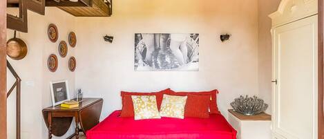 A bedroom with a red bedspread, a wooden desk with a chair, a framed picture on the wall, and a staircase.