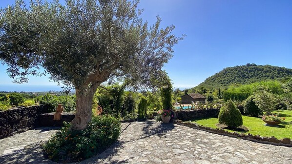 A stone pathway leading to a house with a view of the sea and a hill in the background.
