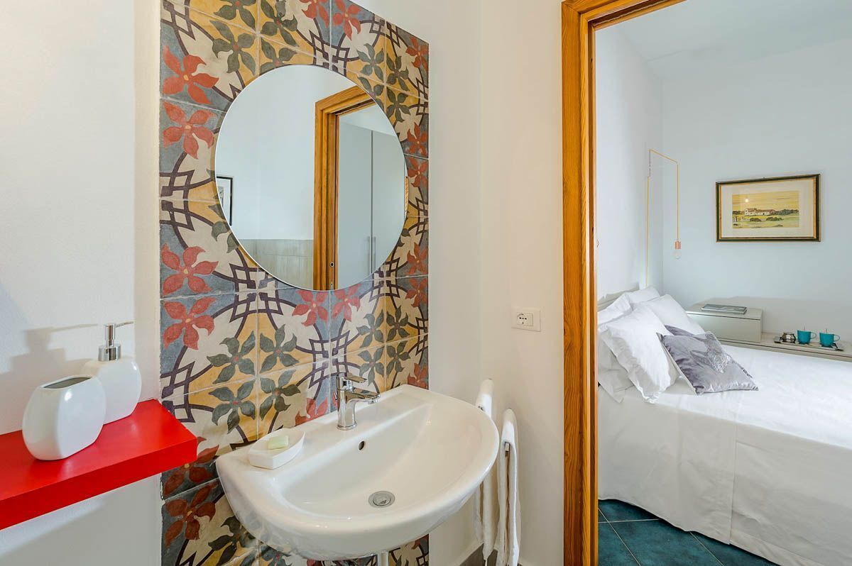 Une salle de bain avec un lavabo blanc, une étagère rouge et un miroir agrémenté d’un pare-feu en mosaïque à motifs floraux.
