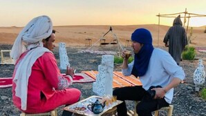 Two people sitting at a table with a teapot and cups, in a desert setting with tents and a cross structure in the background.