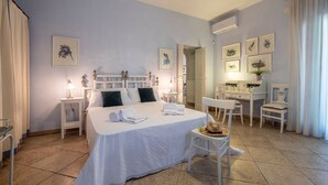 Habitación de hotel con una cama grande, mesitas de noche, un escritorio y sillas. Se observan cuadros enmarcados en las paredes y un candelabro sobre la cama.