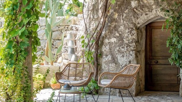 Un espacio de comedor al aire libre rústico con muros de piedra, sillas de mimbre y una mesa servida con frutas y té.