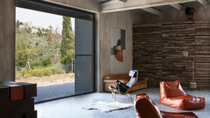Ein modernes Interieur mit einer großen Schiebeglastür, die zu einem schönen Ausblick ins Freie führt, einem zeitgenössischen Kunstwerk an der Wand und einer Mischung an Sitzgelegenheiten, darunter ein Ledersofa und Sitzsäcke.