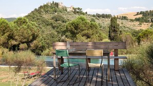 Eine Holzterrasse mit Bank und Stühlen, die auf einen Pool mit einem Hügel und Ruinen im Hintergrund blickt.
