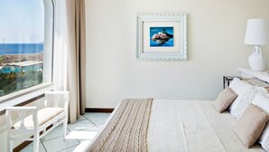 Une chambre avec un grand lit, une chaise, un tableau encadré au mur et une vue sur la mer par la fenêtre.